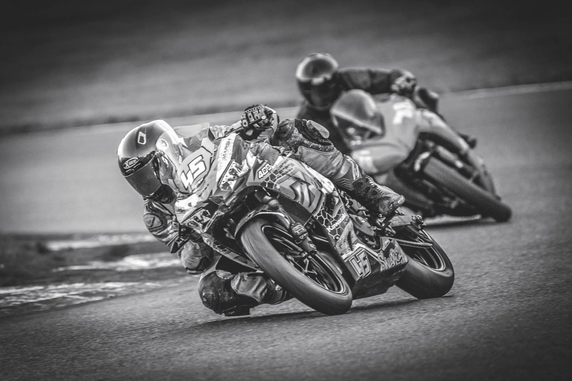 Two motorcycle racers leaning into a turn on a race track, black and white photo.