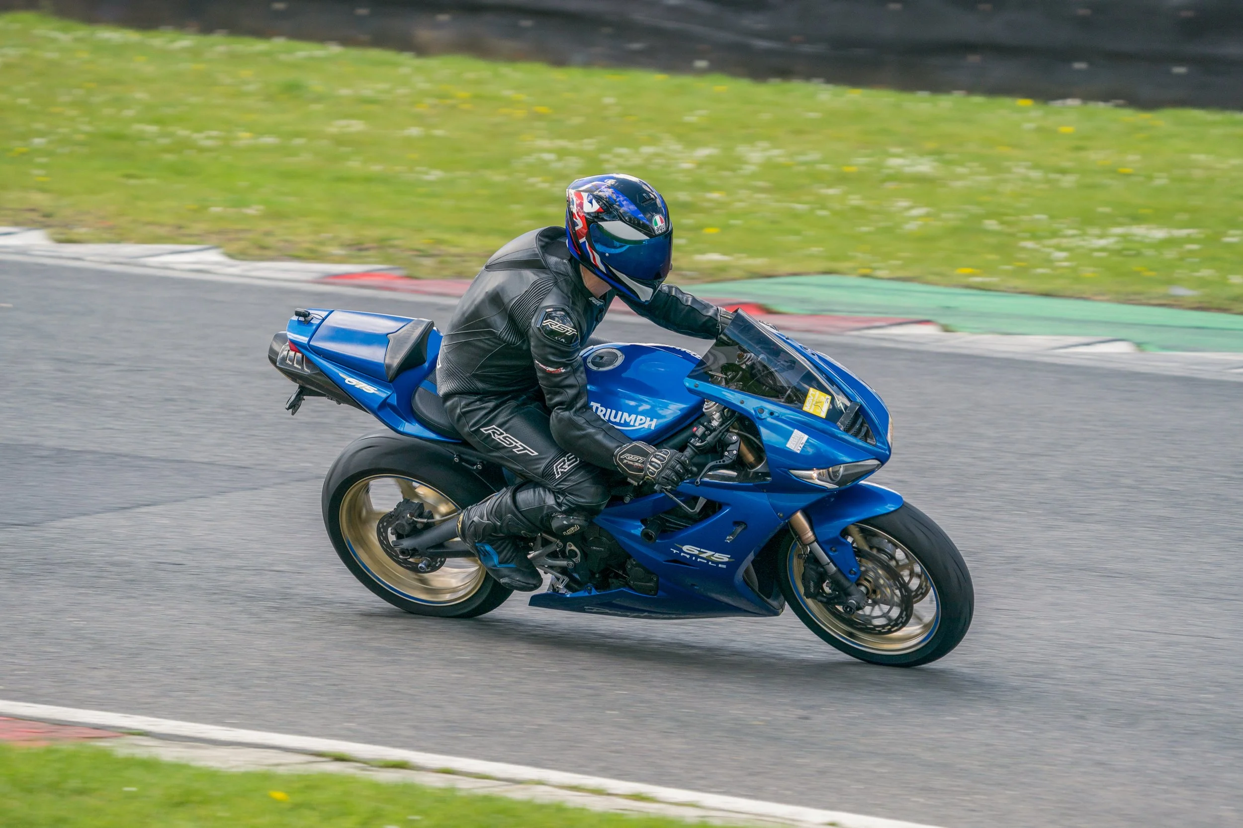 Motorcyclist wearing black gear and a blue helmet riding a blue Triumph racing motorcycle on a curved track.