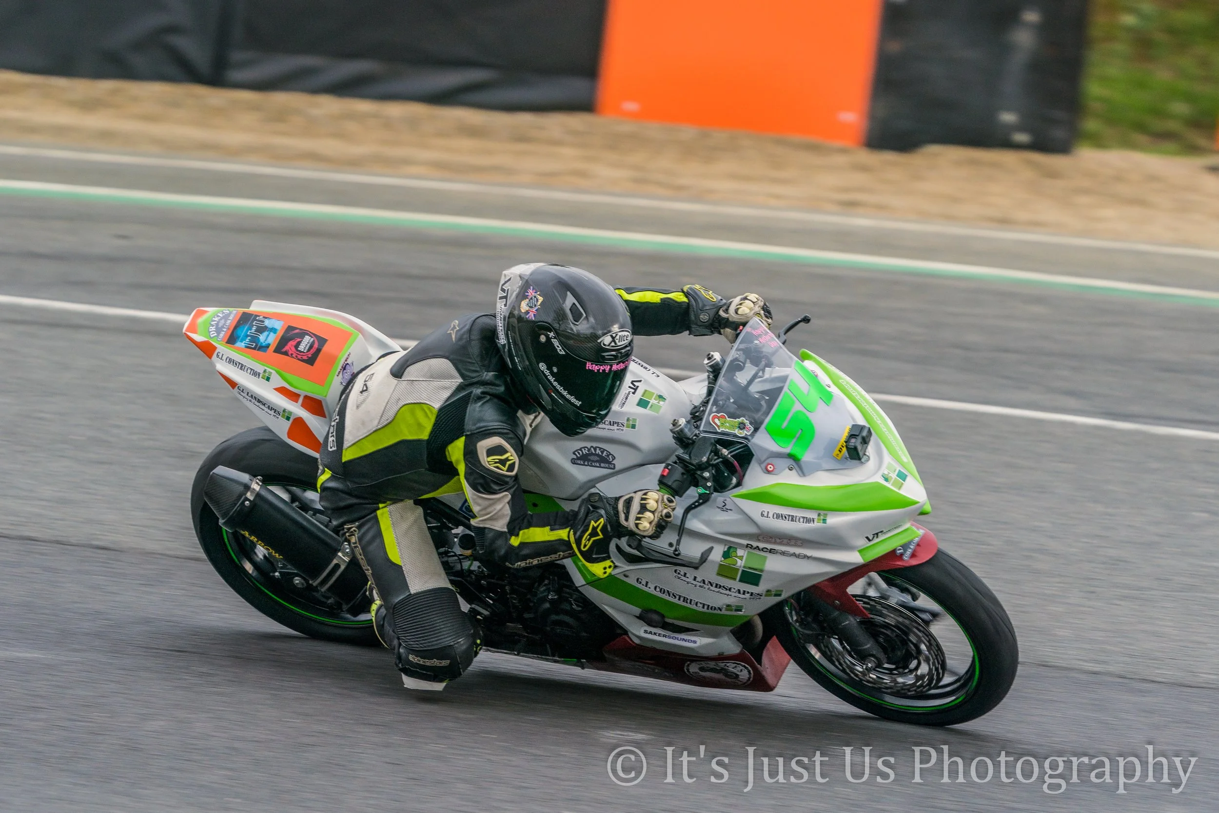Motorcycle racer leaning into a curve on a racetrack, wearing black and gray racing gear with yellow accents, black helmet, and green and white motorcycle with various sponsor stickers.