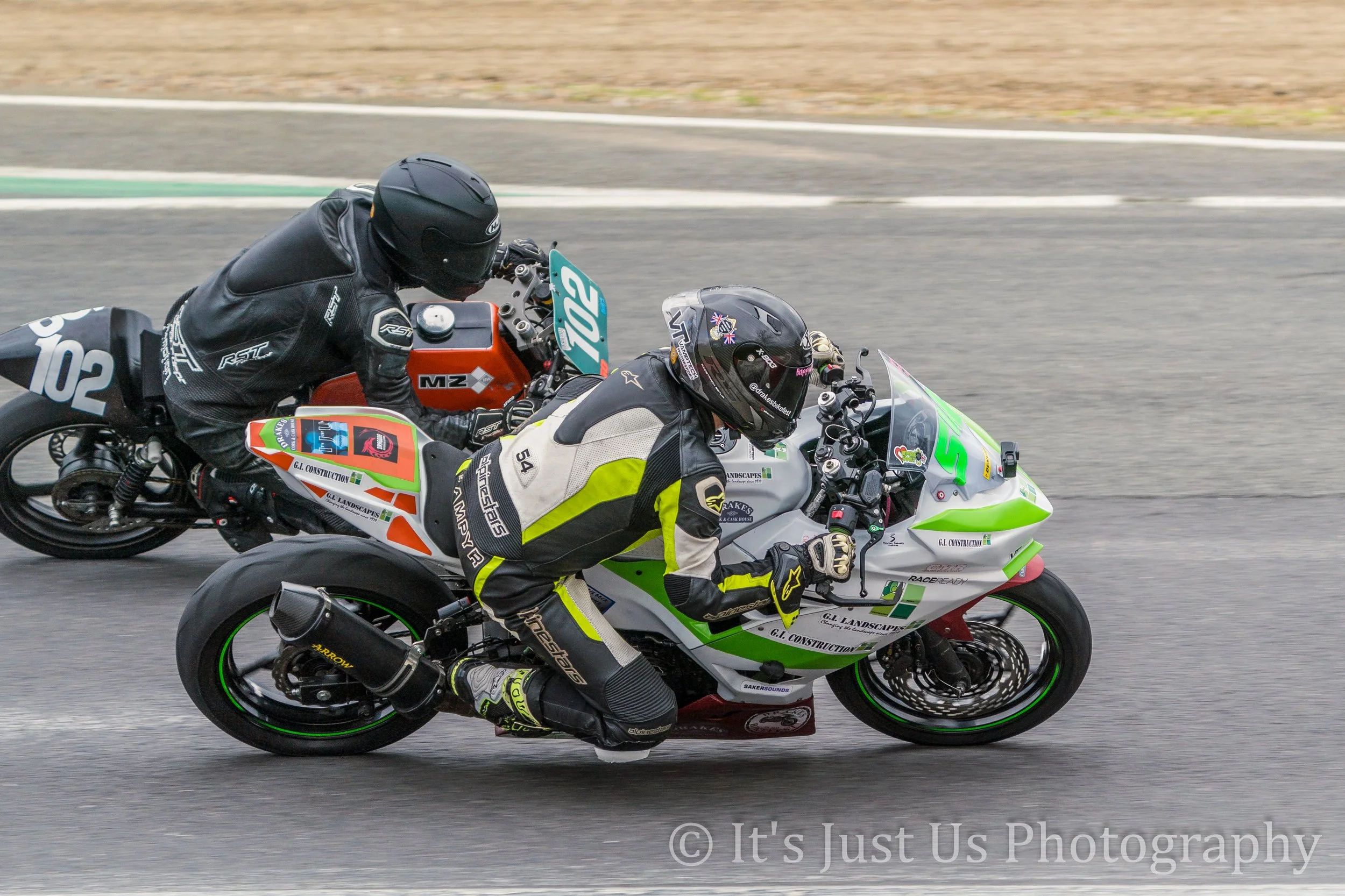 Two motorcycle racers leaning into a turn on a racetrack, wearing full racing gear and helmets, with a blurred asphalt background.