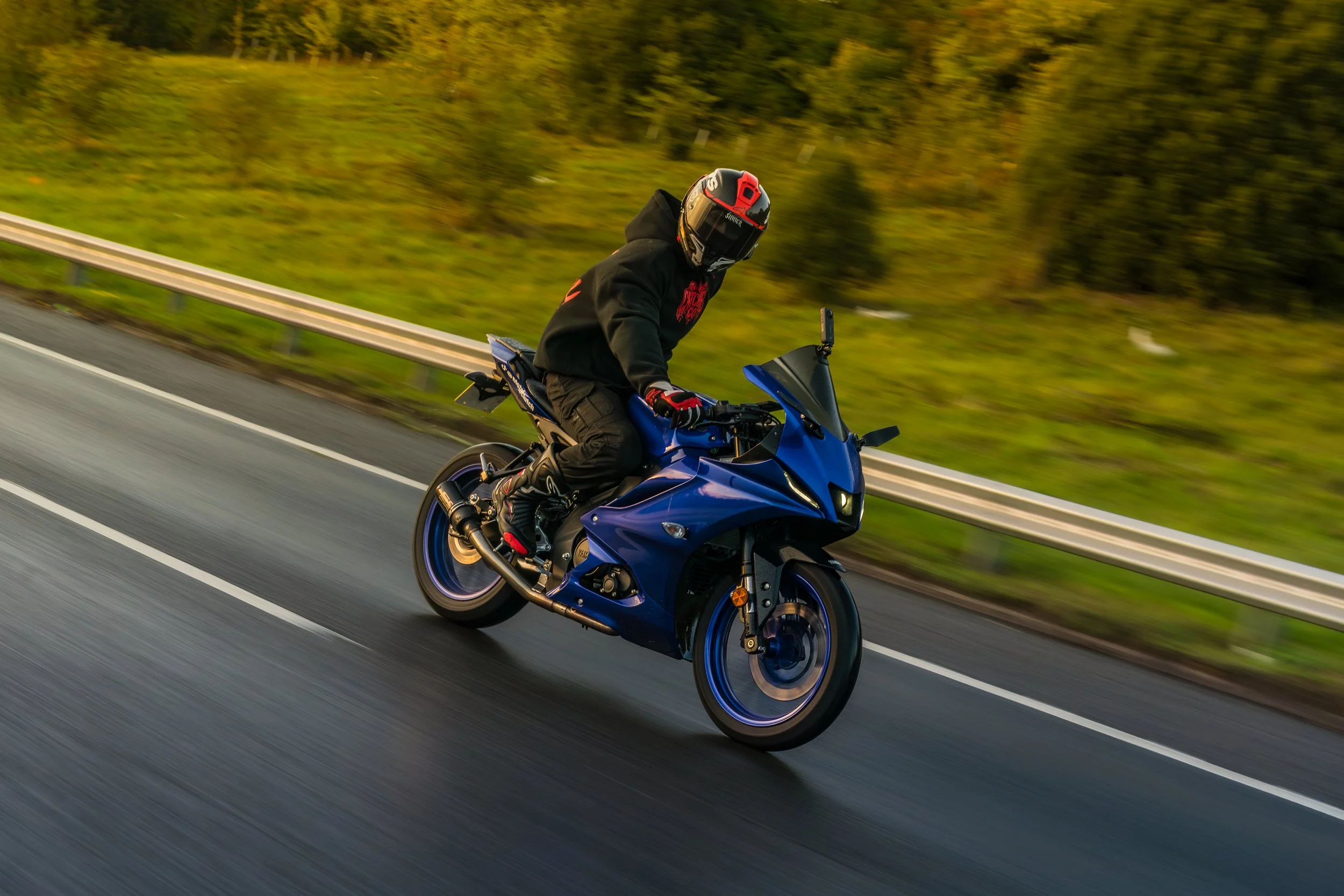 A person riding a blue motorcycle on a highway during daytime. The rider is wearing a black hoodie, black pants, gloves, and a black helmet with red accents. The highway has white lane markers, and the background includes a grassy area and trees.
