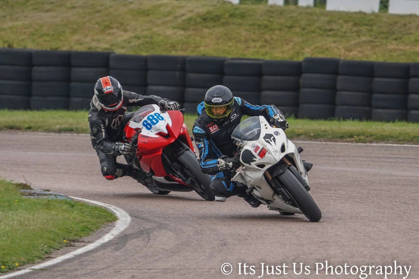 Two motorcycle racers leaning into a turn on a race track, one on a red bike with the number 888 and the other on a white bike with the number 7, with a black tire barrier and grassy area in the background.