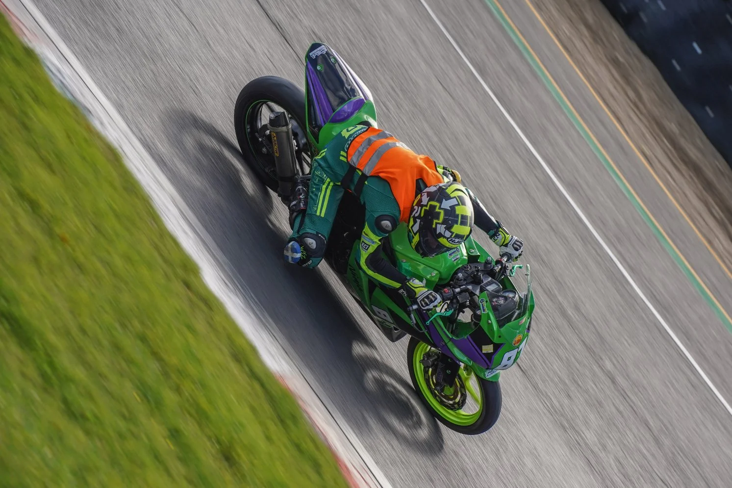 Motorcycle racer in green and purple suit leaning into a turn on a race track, with the motorcycle matching in green and purple, on a cloudy day.
