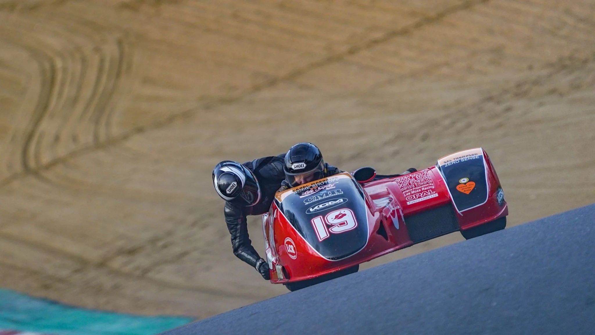 A person in racing gear riding a red sidecar motorcycle on a racetrack with a tan dirt background.