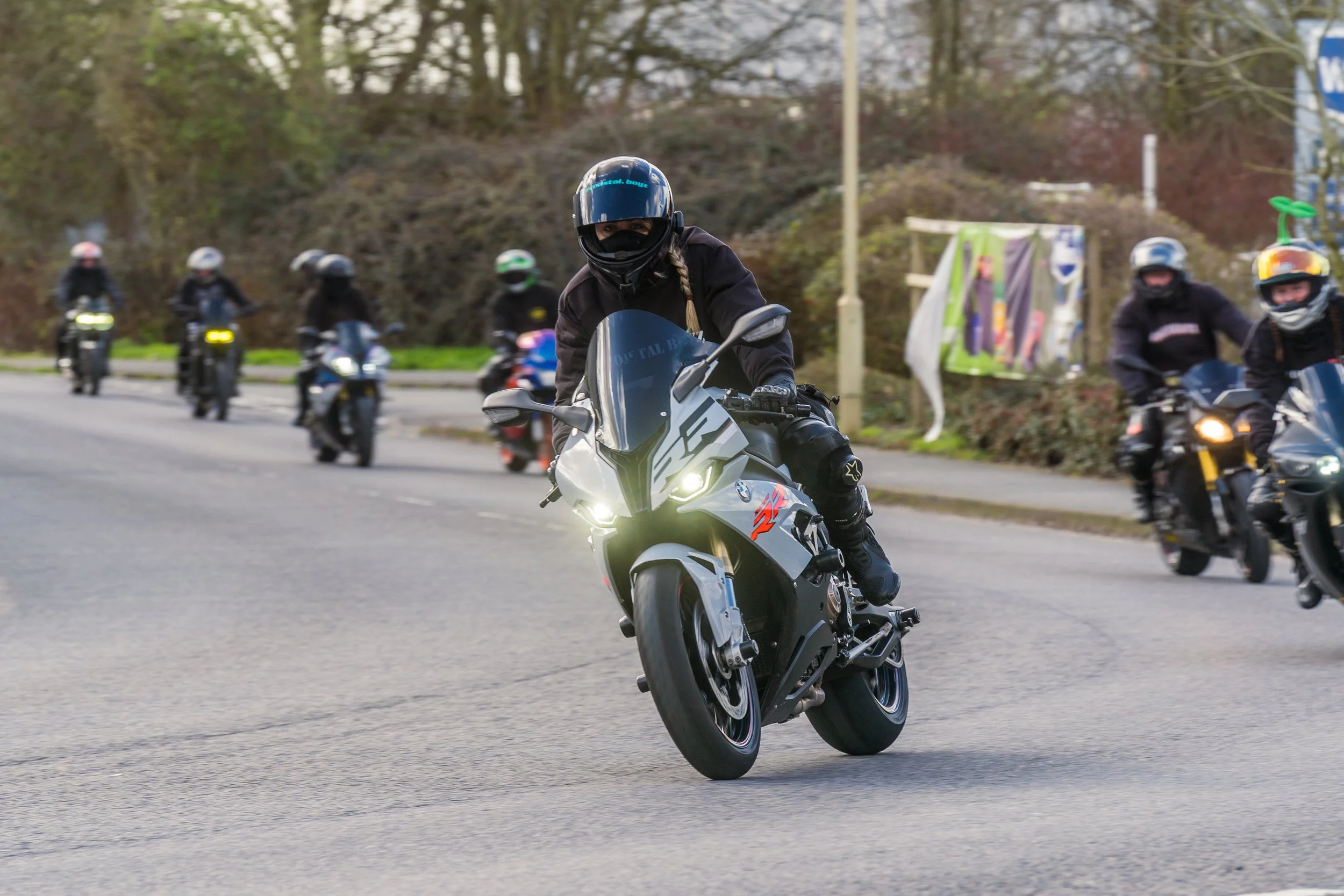 A group of motorcyclists riding on a road, with the rider in front leaning into a turn. The riders are wearing helmets and protective gear, and the background features trees and street signs.