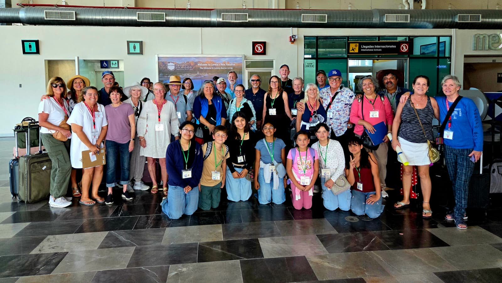 A large group of people, including children and adults, poses for a photo at an airport terminal in front of the arrivals area. Some people are holding luggage, and many are wearing lanyards and identification badges. There are signs for international arrivals in the background.