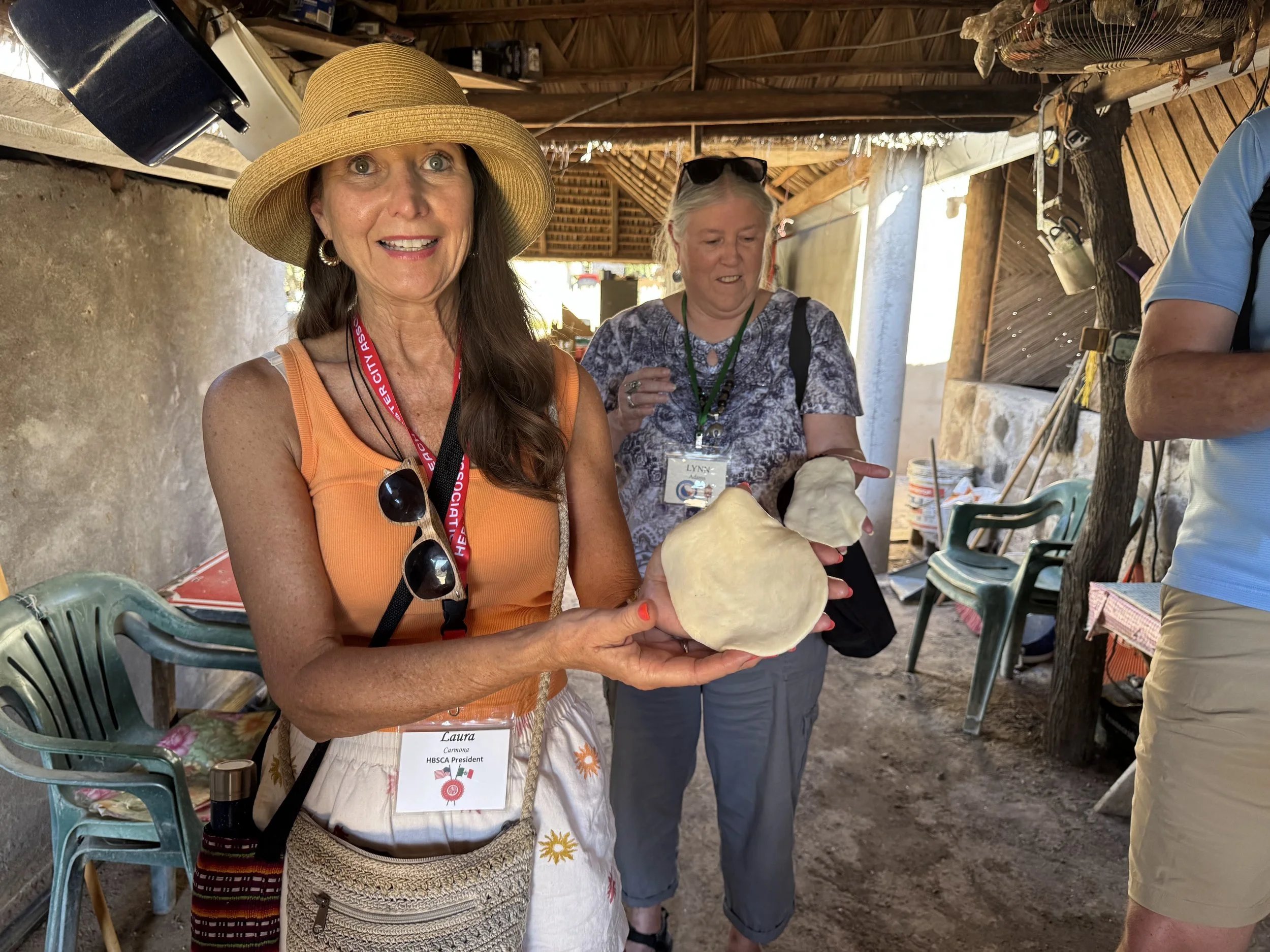 A woman wearing a straw hat, orange top, and sunglasses hanging from her neck, smiling and holding a large shell in an outdoor rustic setting. Another woman nearby is also holding a shell, with chairs, tools, and a tree in the background.