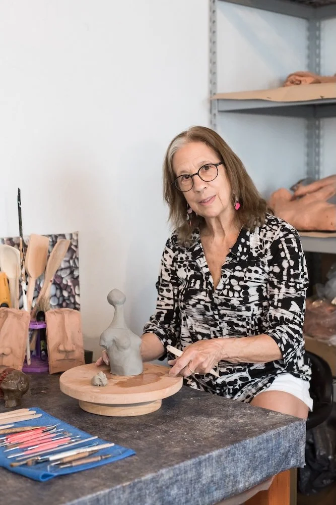 An older woman with glasses and earrings working on a sculpture in a studio, surrounded by clay tools and sculptures.
