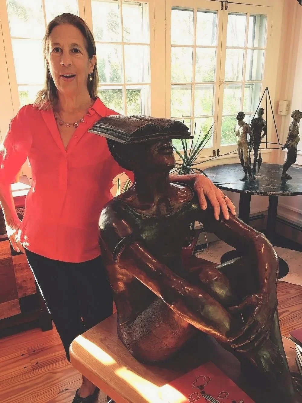 A woman in a red shirt standing next to a large wooden sculpture of a woman with a book on her head, in a room with large windows and art sculptures in the background.