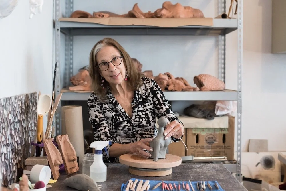An older woman with glasses and earrings shaping clay on a pottery wheel in her studio, with shelves of clay sculptures in the background.