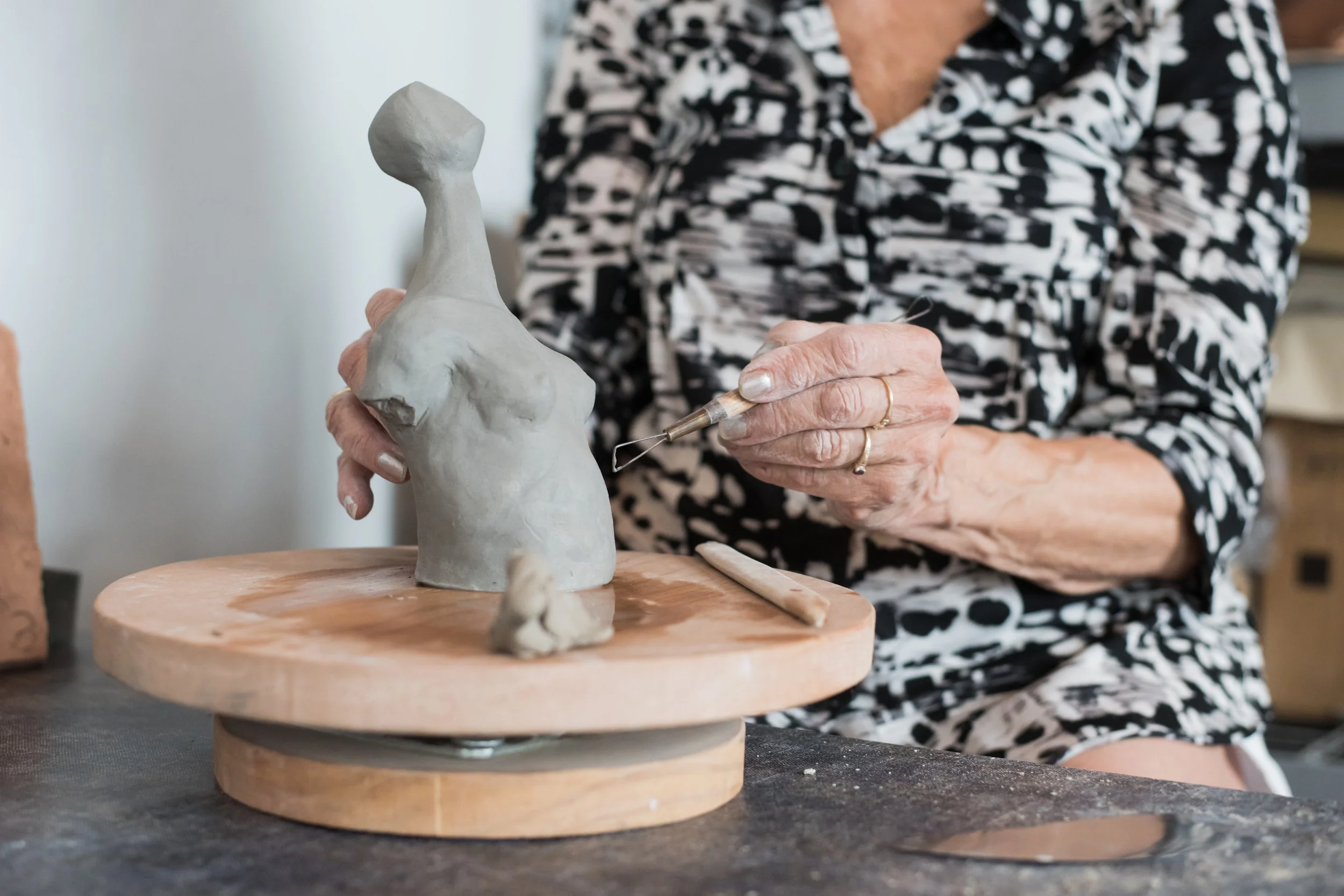 A person is shaping a ceramic sculpture of a torso with a raised arm using a tool, sitting at a workstation. The sculpture is on a wooden turntable, and the person wears a black and white patterned shirt.