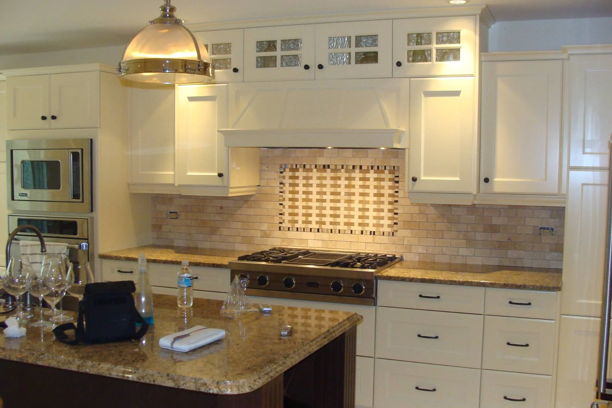 A kitchen with cream-colored cabinets, granite countertops, a gas stove, a tiled backsplash, and a large island with wine glasses and utensils on it.