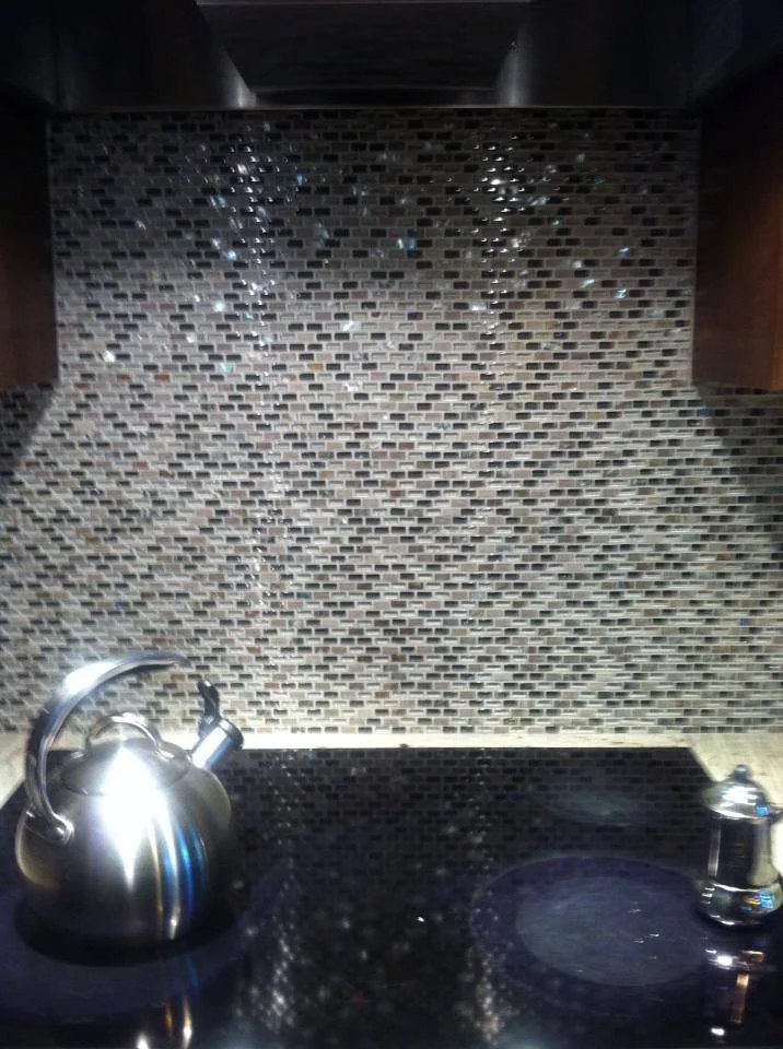 A kitchen stove with a kettle on the left and a small object on the right, featuring a mosaic tile backsplash with water droplets.
