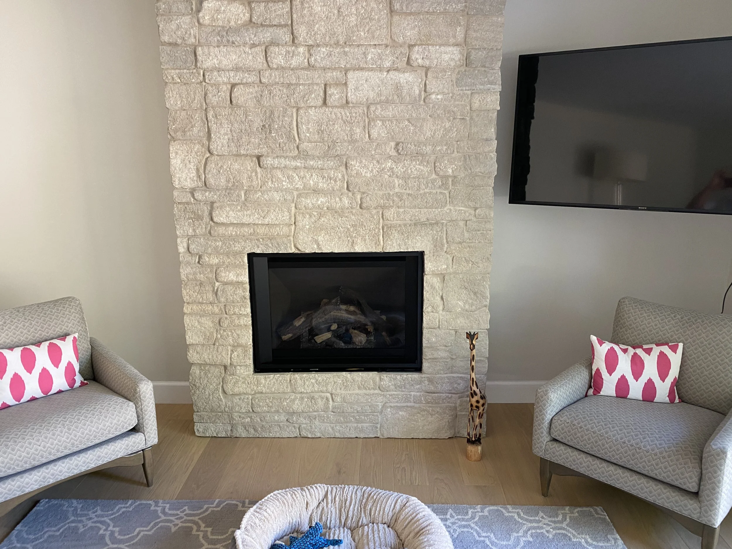 Living room with a beige stone fireplace, a wall-mounted flat screen TV, two upholstered armchairs with pink and white patterned pillows, a giraffe figurine on the floor, a woven basket with blue toys, and a patterned area rug.