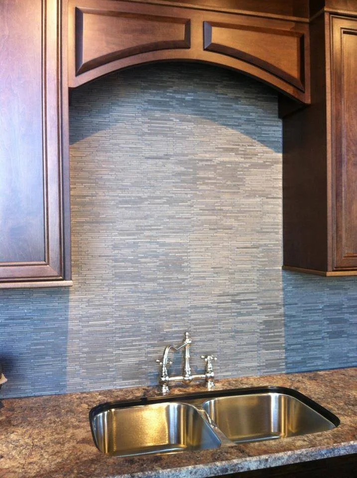 Kitchen sink with dual basins, chrome faucet, and a brown speckled countertop against a gray tile backsplash with dark wood cabinets above.