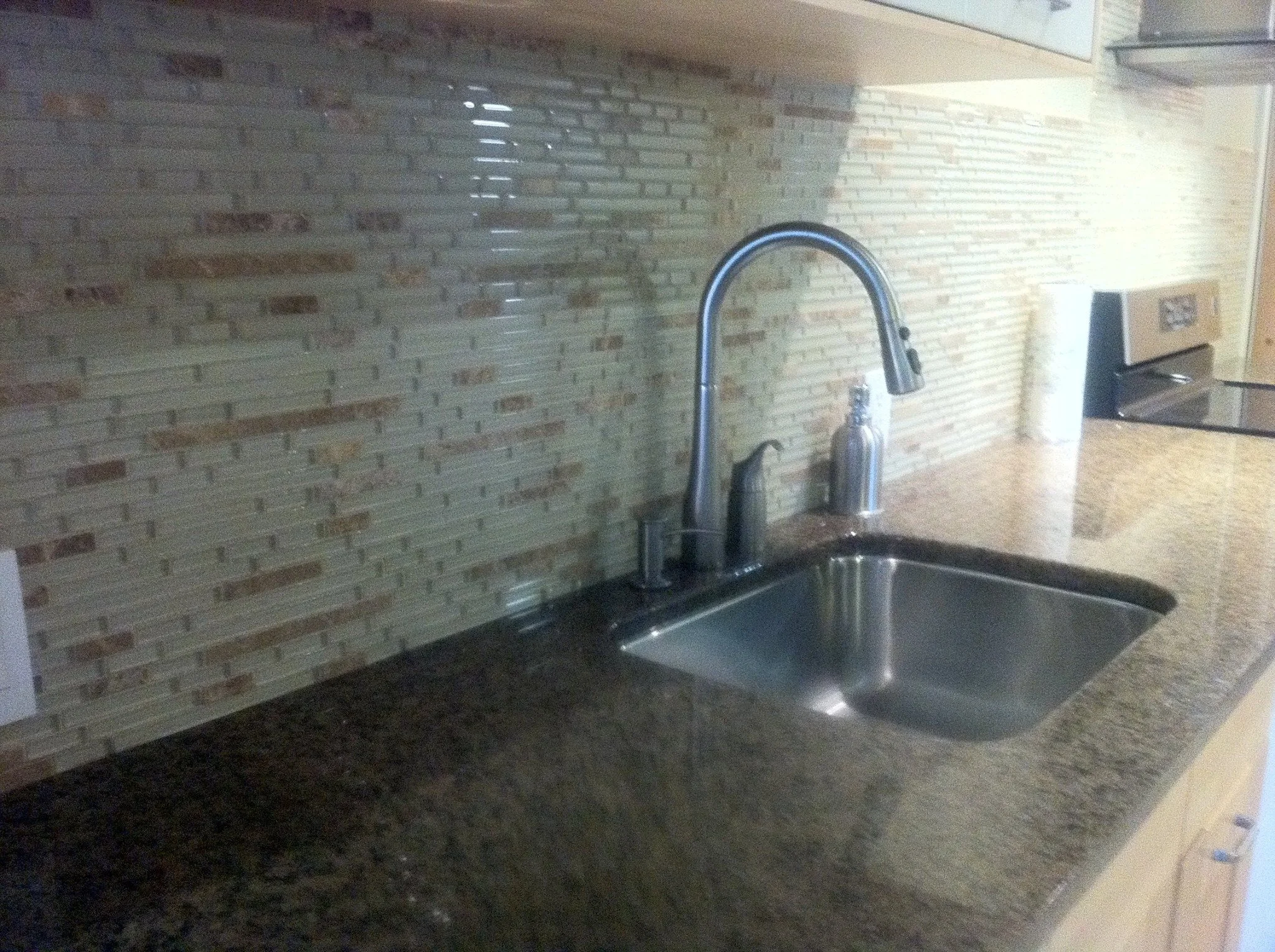 Kitchen countertop with a stainless steel sink, modern faucet, soap dispenser, paper towel roll, and stove in the background. The backsplash is made of rectangular tiles in beige and brown tones.