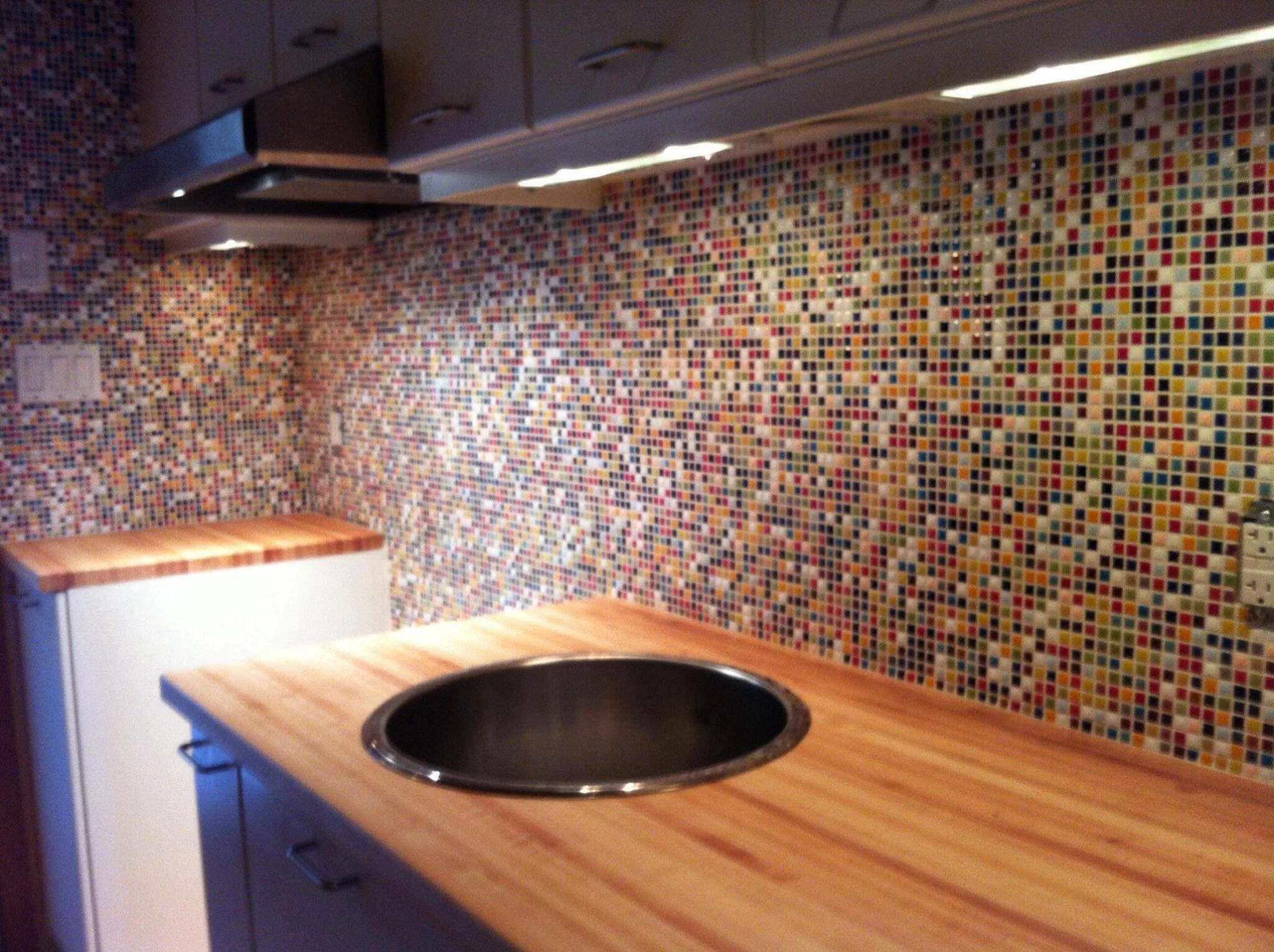 A kitchen with a mosaic tile backsplash, a wooden countertop, and a circular sink, with a white cabinet in the corner.