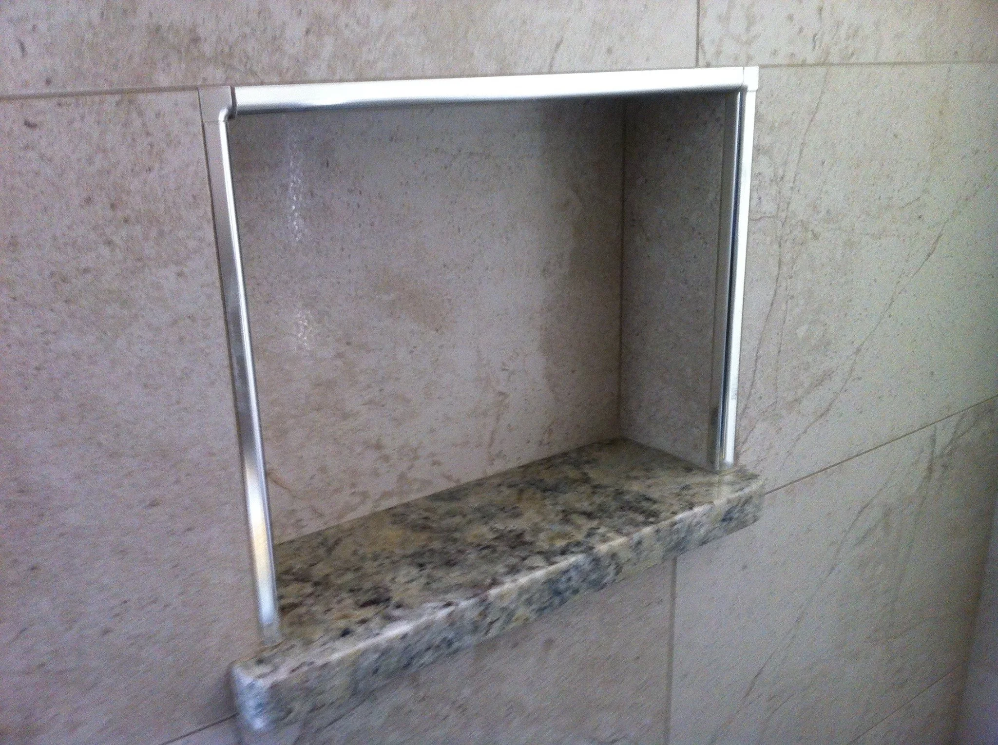A recessed niche in a beige tile wall with a granite shelf and a metallic frame.