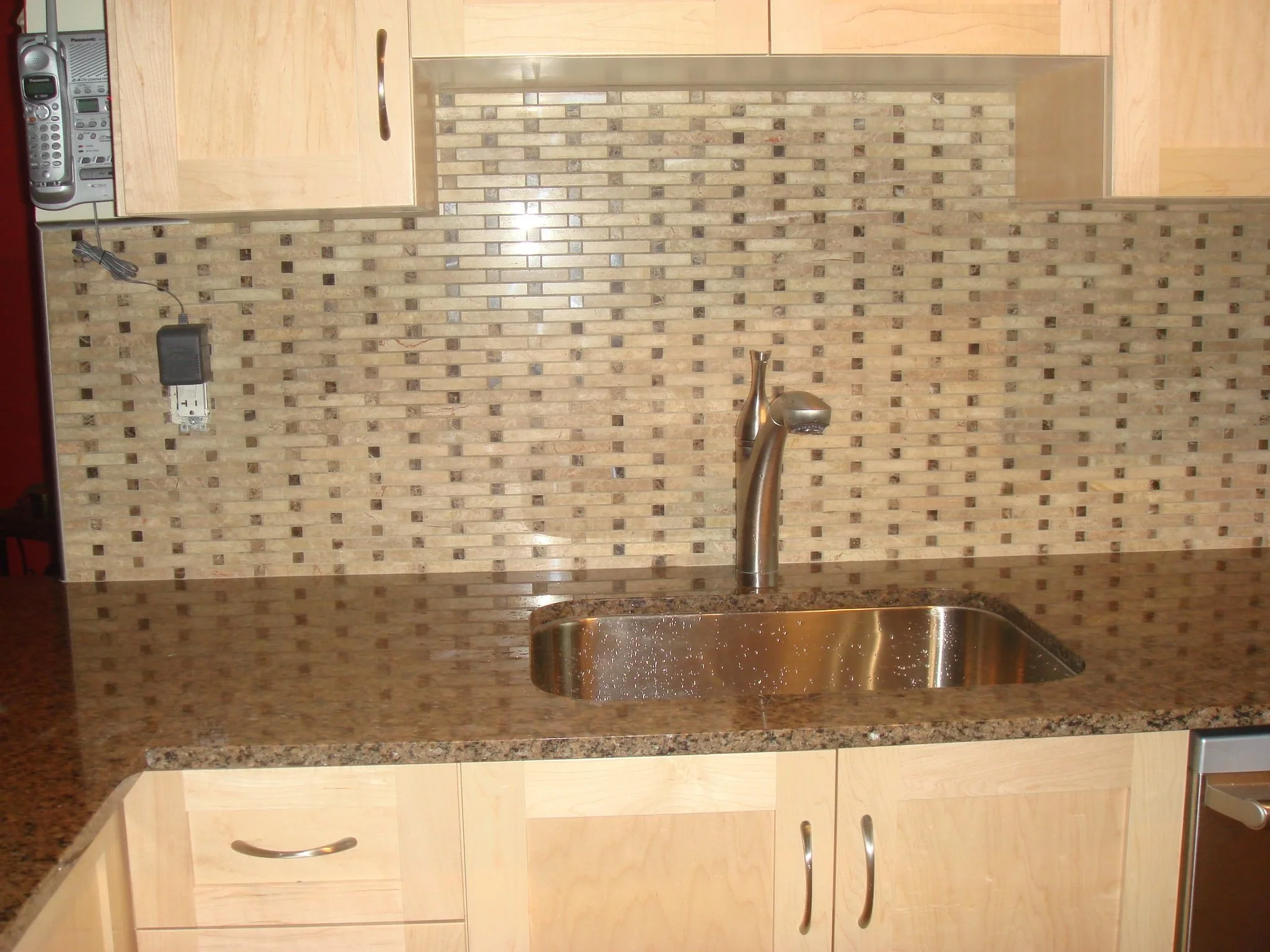 Kitchen countertop with a stainless steel sink, faucet, light-colored wooden cabinets, and a mosaic tile backsplash.