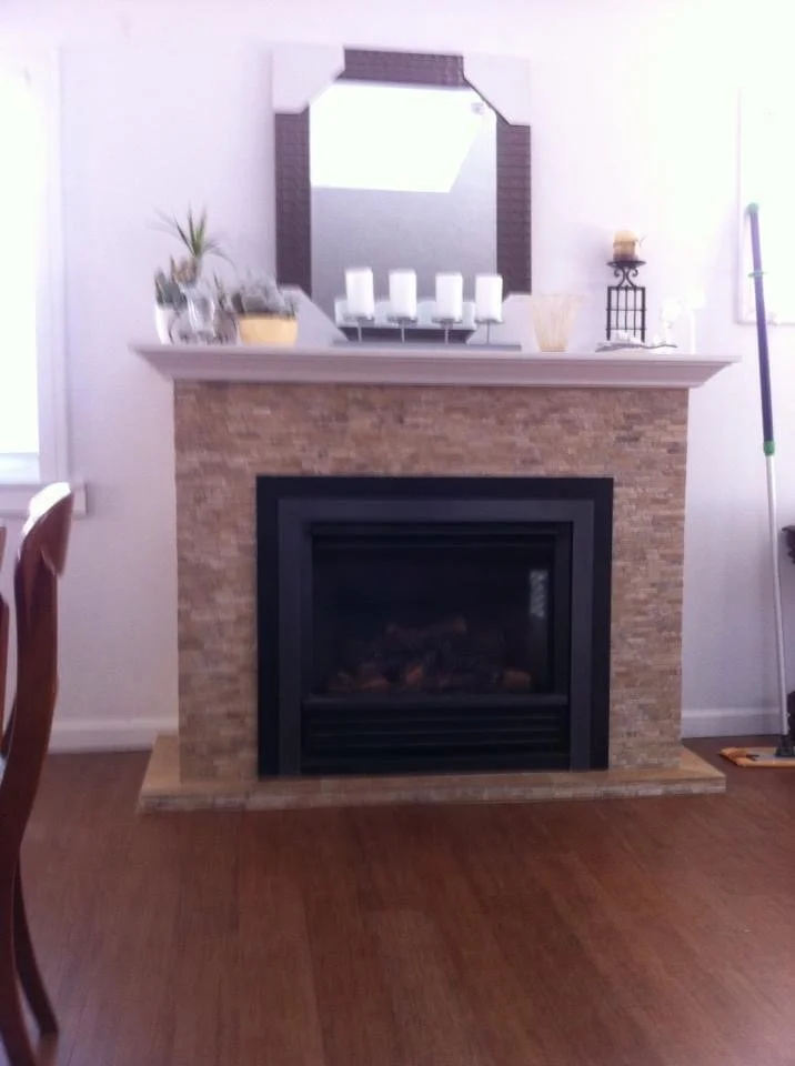 Living room fireplace with a beige stone surround, black frame, a white mantel decorated with candles, plants, and decorative items, and a large mirror above.