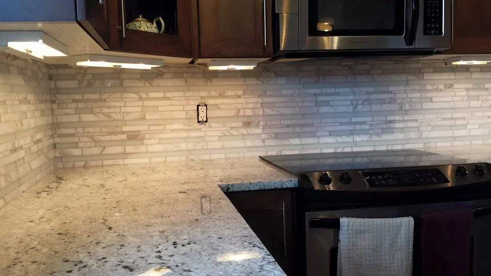 Kitchen countertop with granite surface, backsplash with small rectangular tiles, photographed near stove and microwave.
