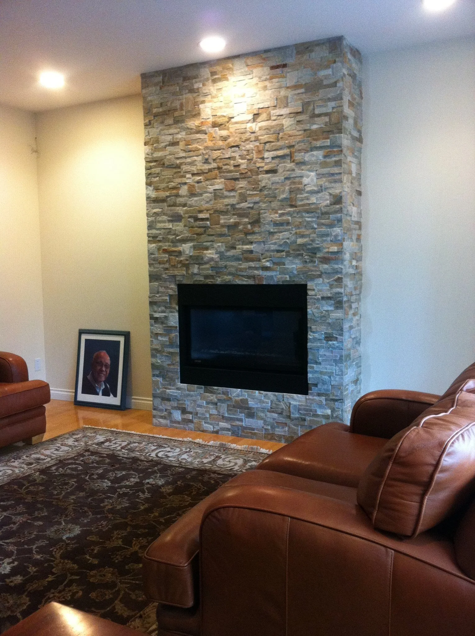 A living room with a stone fireplace at the center, two leather couches, a framed portrait on the floor, a decorative rug, and recessed ceiling lights.