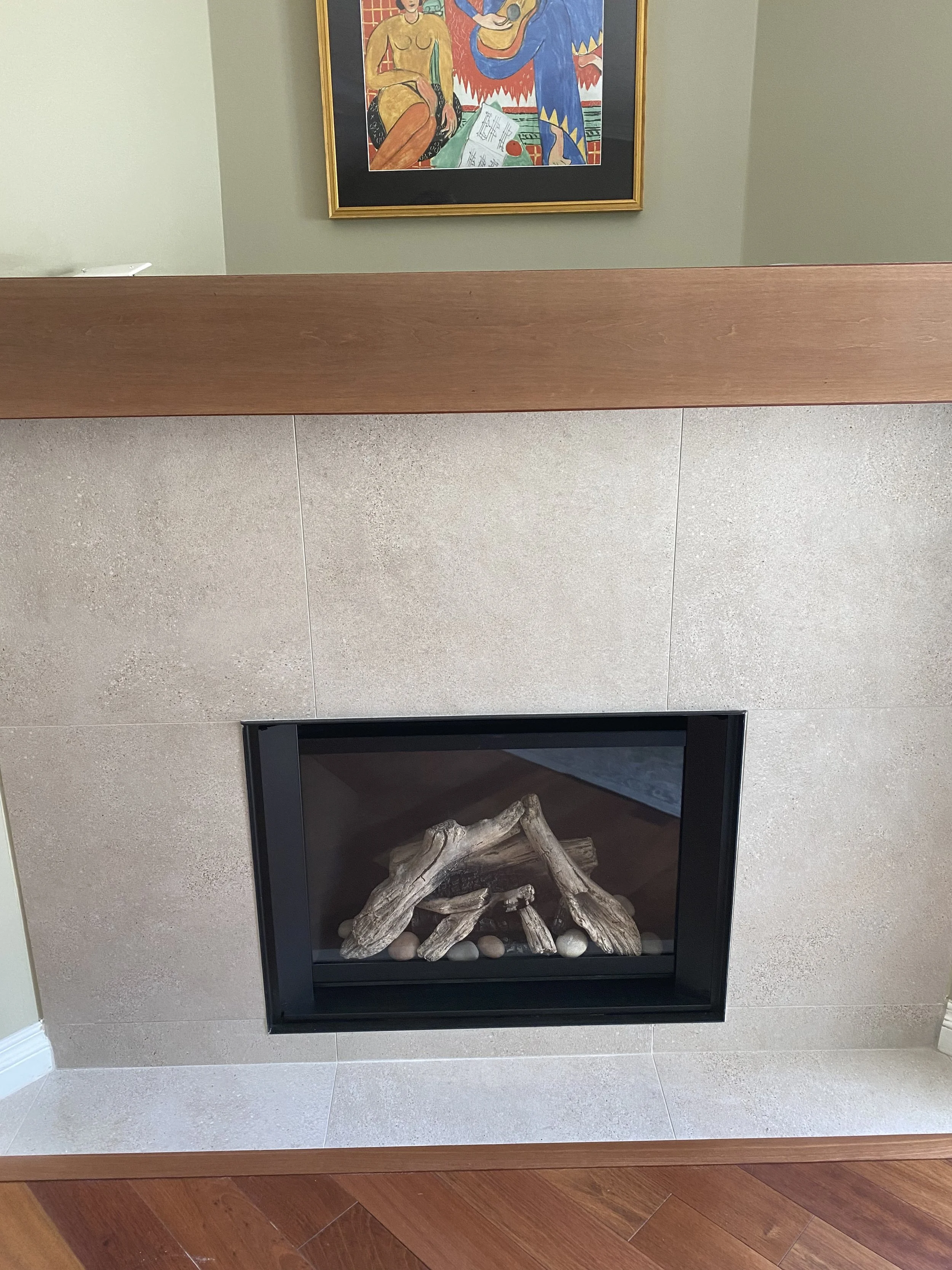 Fireplace with stacked-looking logs and smooth stones inside a beige tiled surround, with a wooden mantel above and an abstract colorful painting on the wall above it.