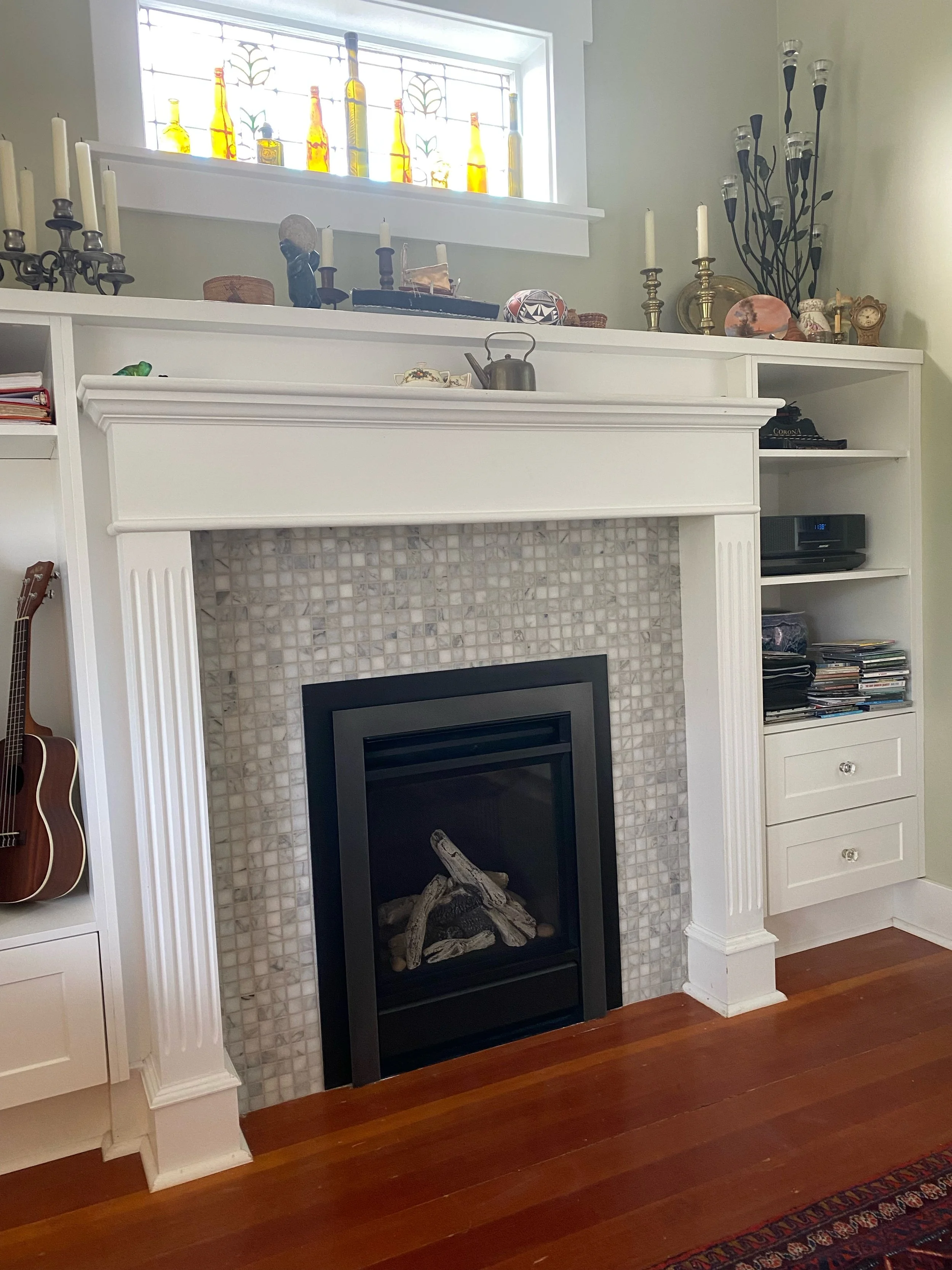 Living room fireplace with white mantel, mosaic tile surround, and artificial logs inside. Above the mantel are various decorative items, including candles, vases, and sculptures. To the left, there is a guitar, and on the right, built-in shelves wit