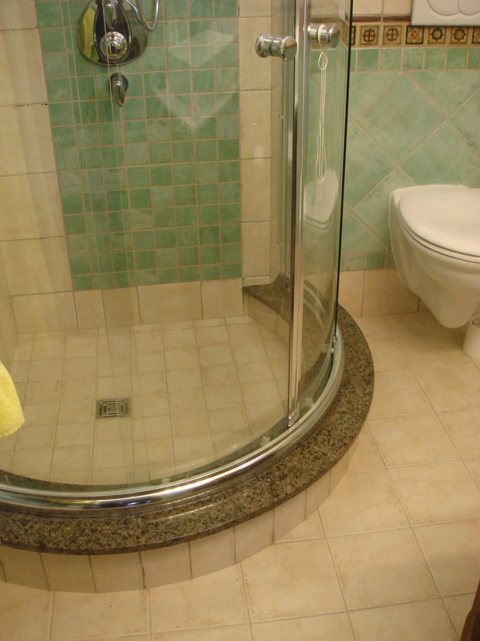 A bathroom with a curved glass shower enclosure with a granite base and a wall-mounted showerhead. The floor and walls are tiled in beige and green tiles. There is a white wall-mounted toilet on the right side.