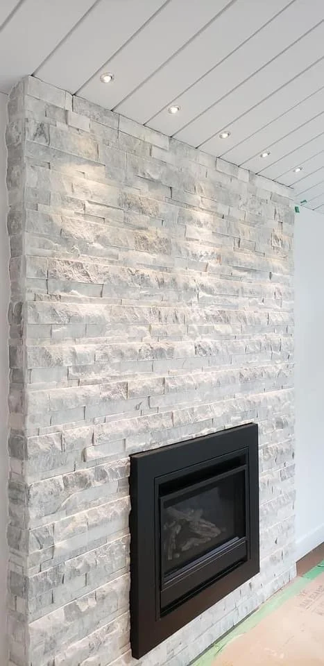 A modern interior fireplace with a black metal frame set into a stone wall made of light-colored bricks, under a white paneled ceiling with ceiling lights.
