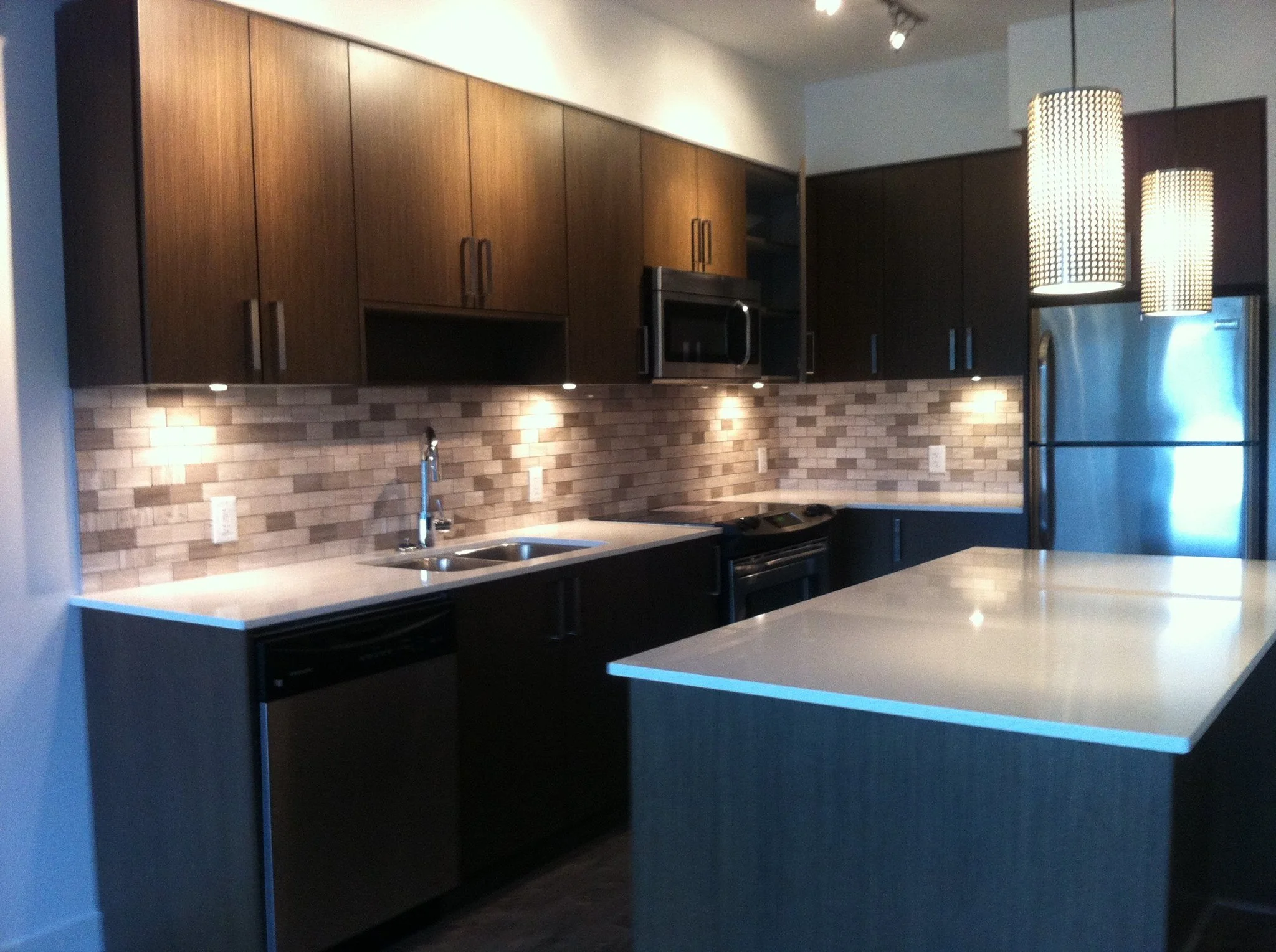 Modern kitchen with dark upper and lower cabinets, white countertops, brick-style beige backsplash, stainless steel refrigerator, oven, microwave, and dishwasher, under-cabinet lighting, hanging pendant lights, and a breakfast island.