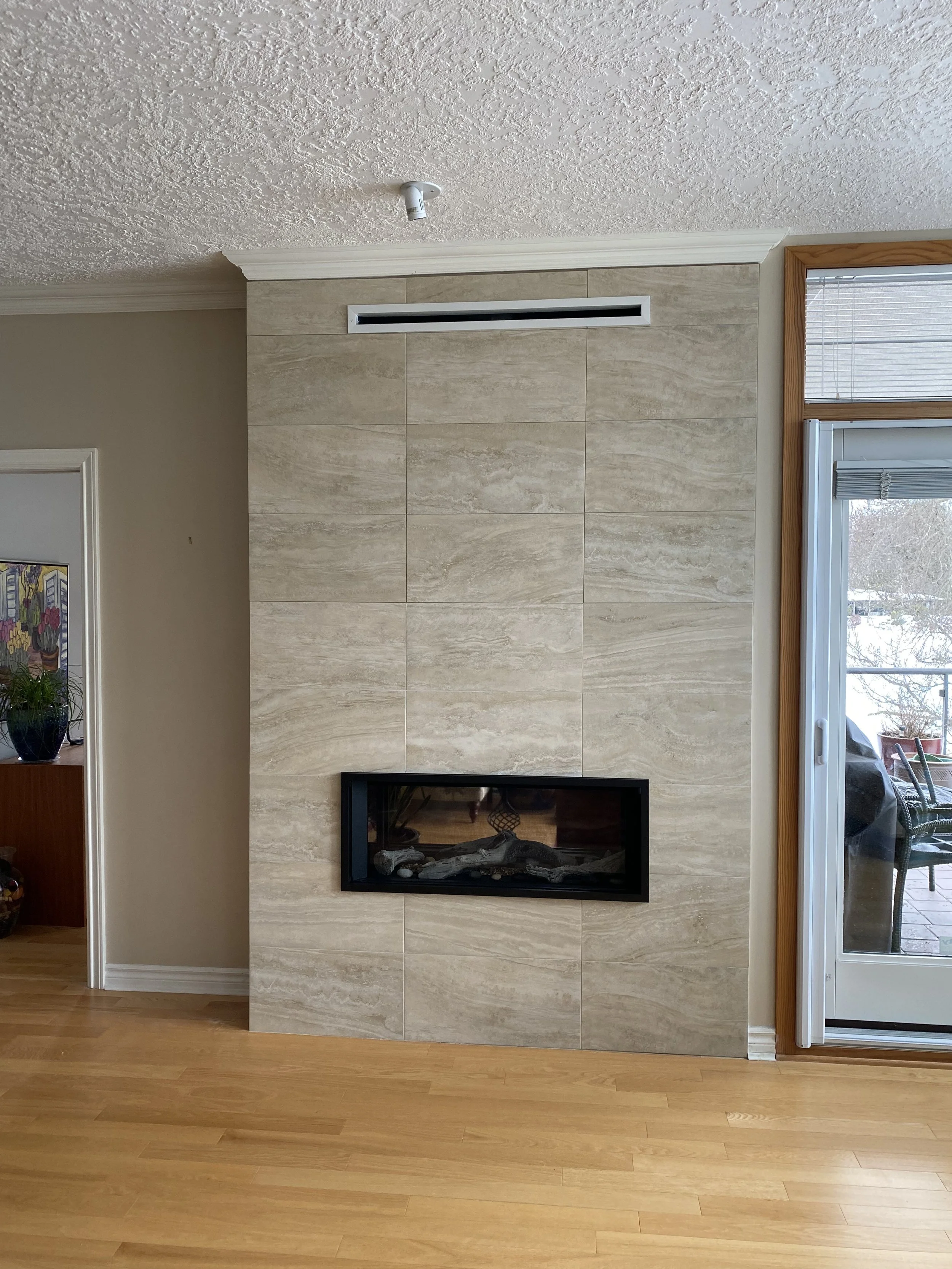 Interior living room with a beige tile wall and a black framed fireplace, wooden floor, and sliding glass door to an outdoor patio.