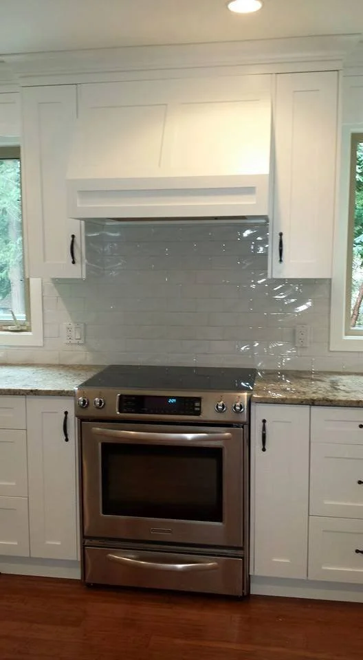 Kitchen with white cabinets, granite countertop, stainless steel oven, gray tile backsplash, and two windows.