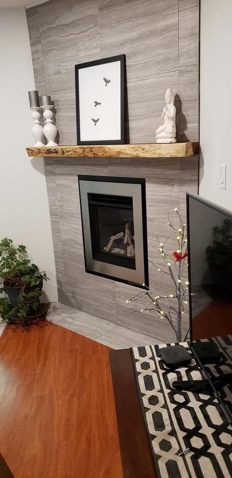 Modern living room corner with a fireplace set into a gray tiled wall, a wooden floating shelf above it with decorative items including a white statue and framed artwork with butterflies, and a potted plant on the left side. There is a small, decorat