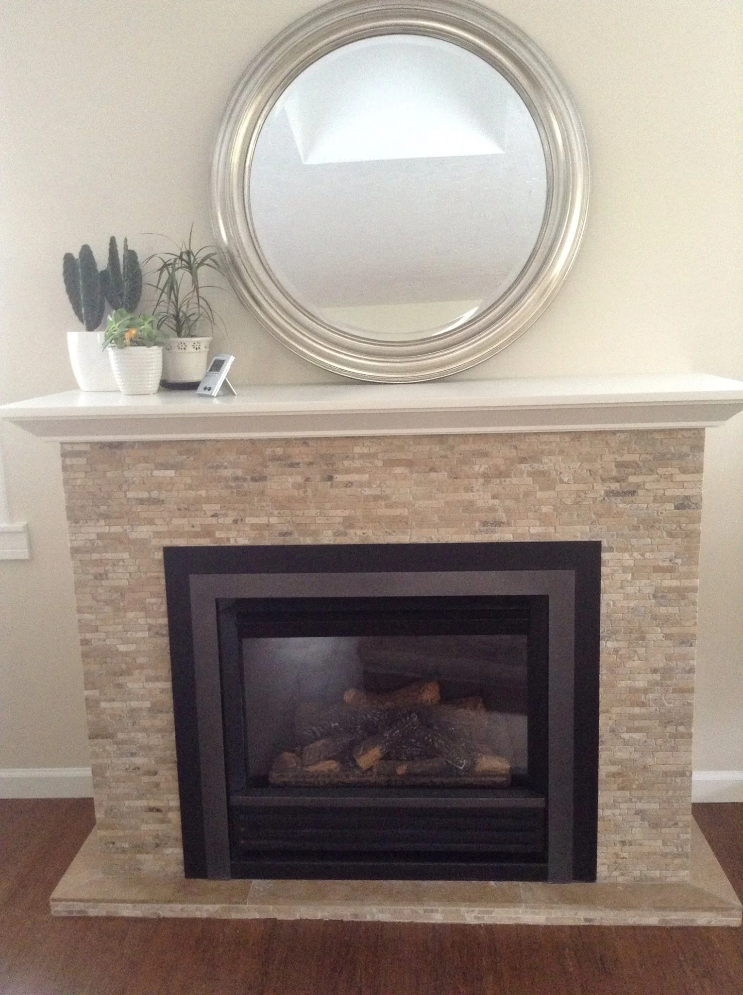 A fireplace with a beige stone surround, black insert, and a white mantel. Above the mantel is a large round silver mirror, and on the left side of the mantel are three potted plants and a cordless phone.