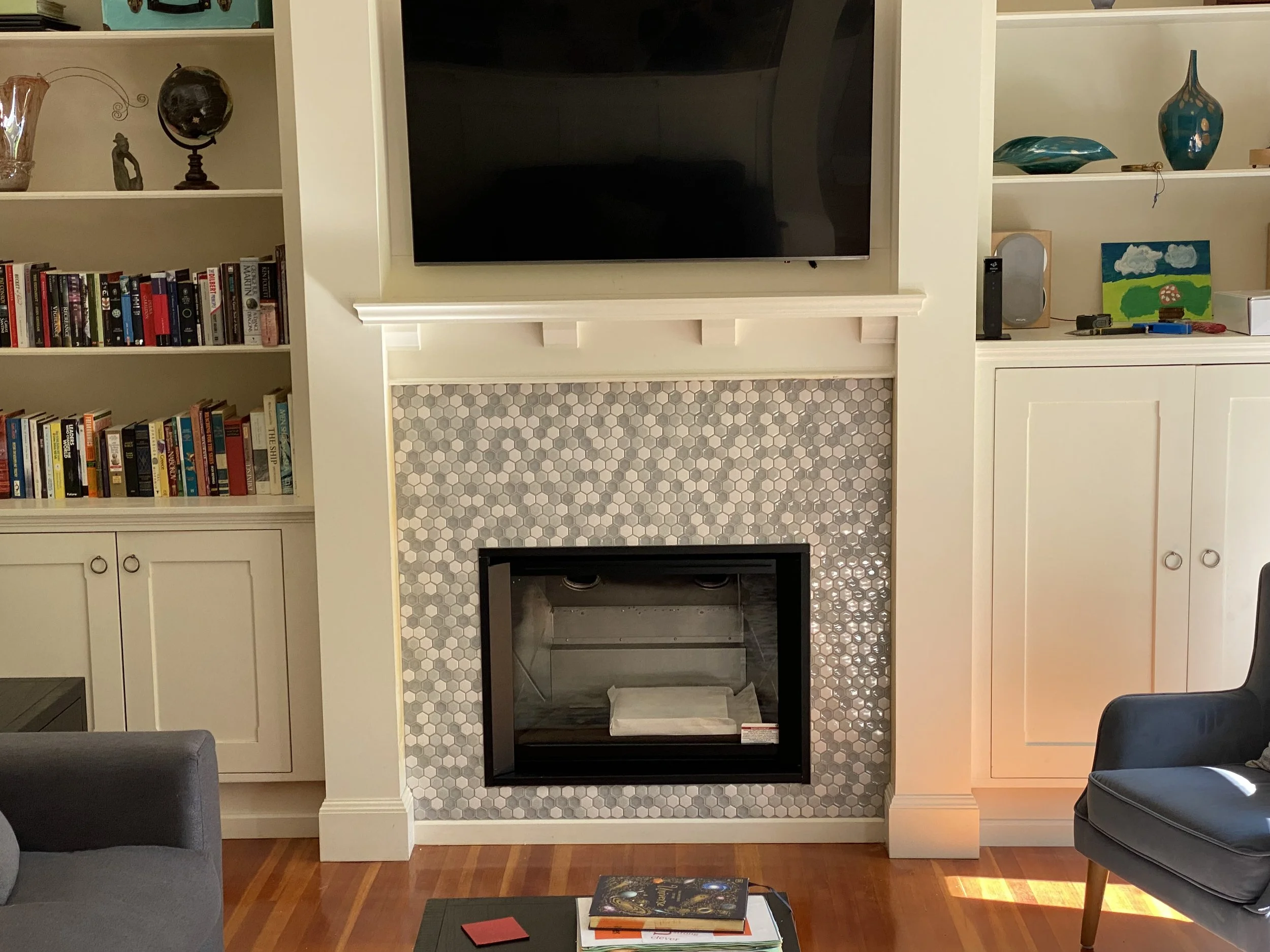 Living room fireplace with a flat-screen TV mounted above, surrounded by white built-in shelves with books, decorative objects, and artwork, with hardwood flooring.