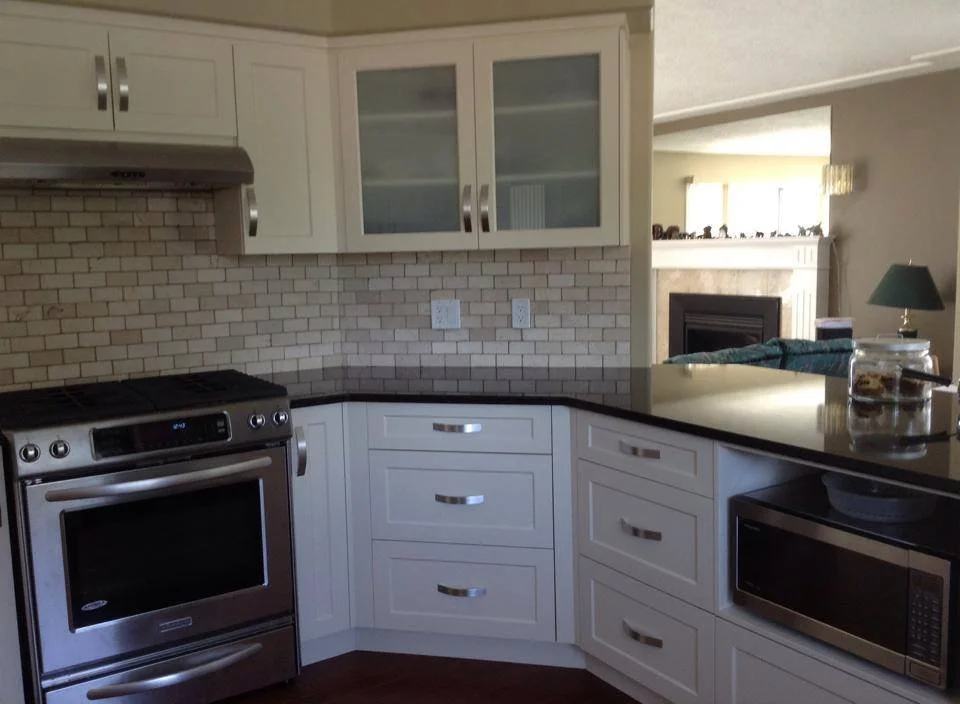 Kitchen with white cabinets, a stainless steel oven, a microwave, and a brick backsplash. Part of a living room with a fireplace and lamp visible in the background.