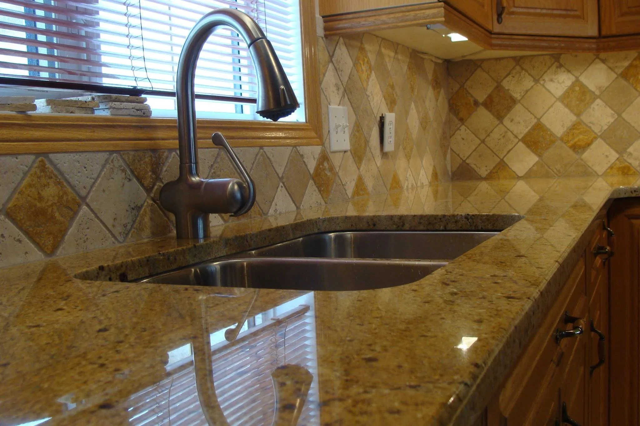Close-up of a kitchen sink with a modern silver faucet, granite countertop, and tiled backsplash featuring beige and brown diamond-patterned tiles. A window with blinds is above the sink, and electrical outlets are to the side.