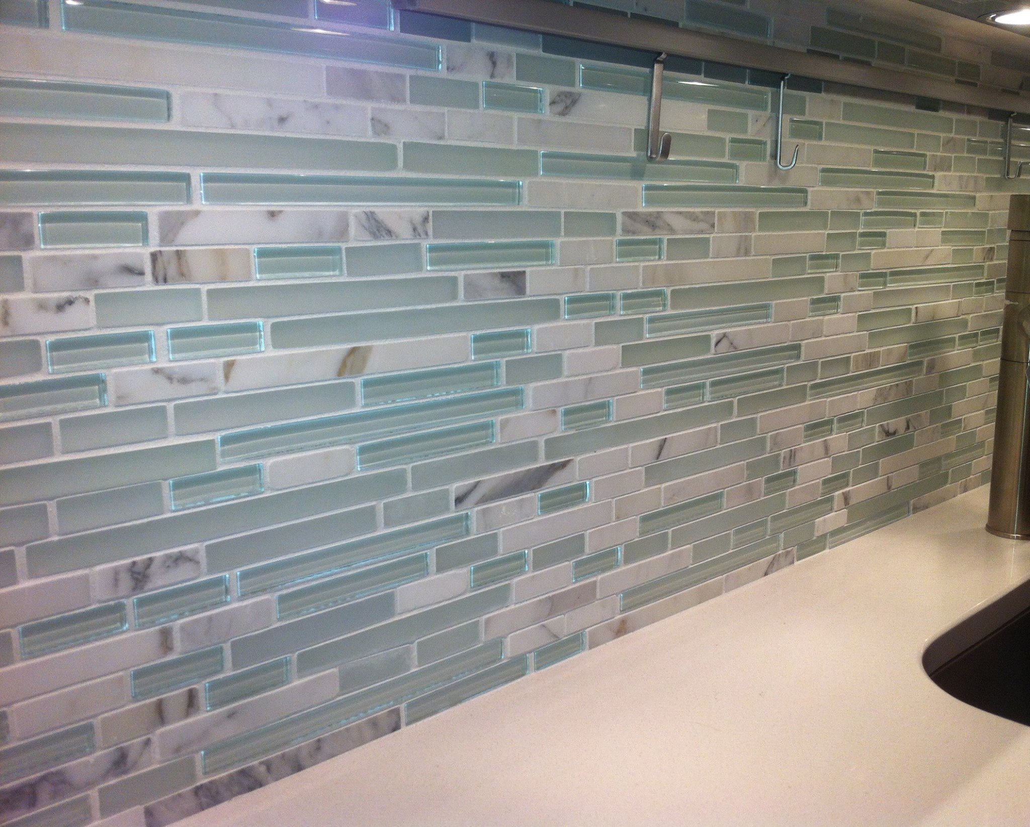 Close-up of a tiled backsplash with a mix of marble, glass, and stone tiles above a white countertop, with metal hooks and part of a sink visible.