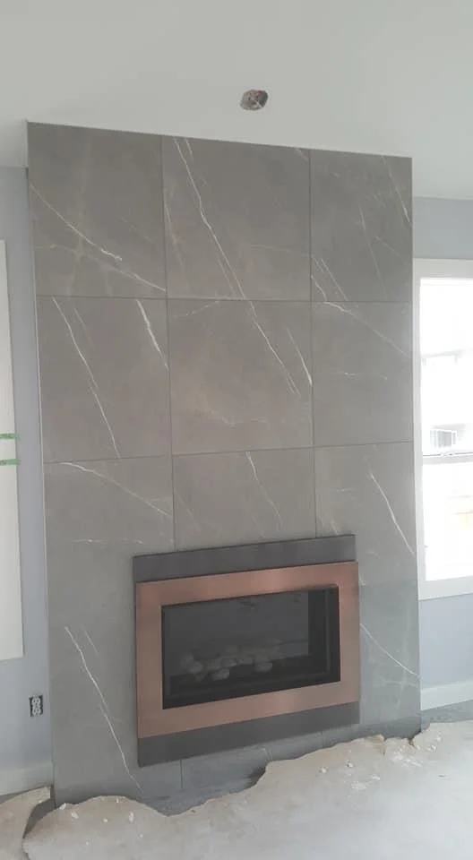 Interior view showing a partially constructed fireplace with gray stone tiles and a metal frame, in a room with unfinished flooring and a window on the right side.