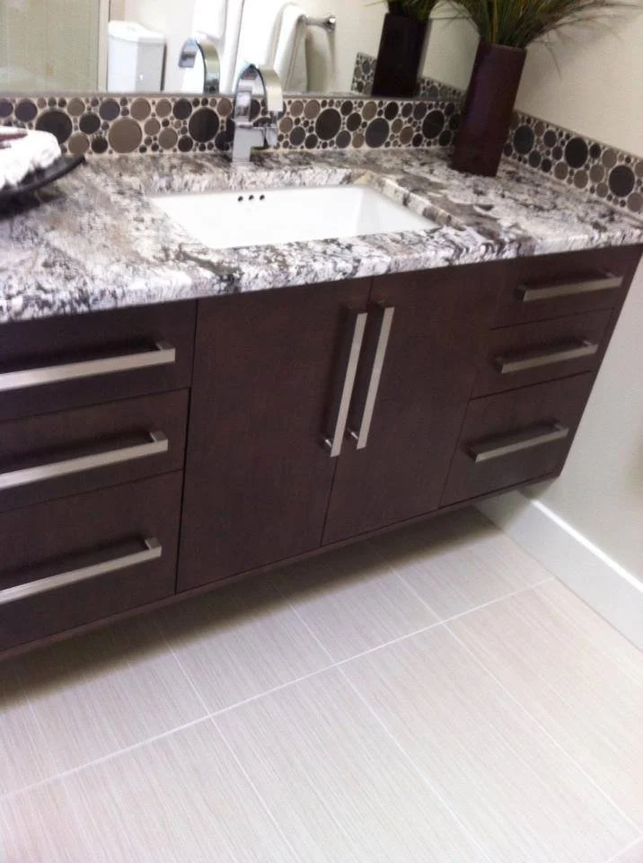 Bathroom vanity with dark wood cabinets, granite countertop, small rectangular undermount sink, and a mosaic backsplash with circular tiles. Two planters with green plants are positioned on the countertop, and a mirror reflects part of the bathroom.