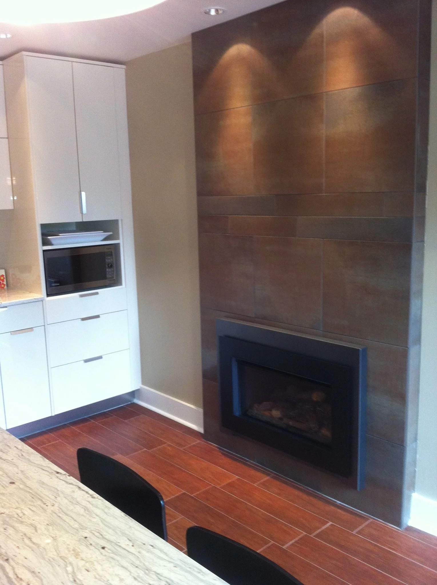 Interior view of a modern kitchen with a gas fireplace, white cabinets, a microwave, a marble countertop, and black chairs.