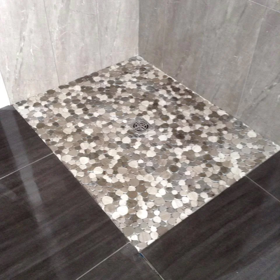 Close-up of a shower stall floor with a drain, featuring small round mosaic tiles in shades of beige, brown, and cream, bordered by larger dark gray tiles.