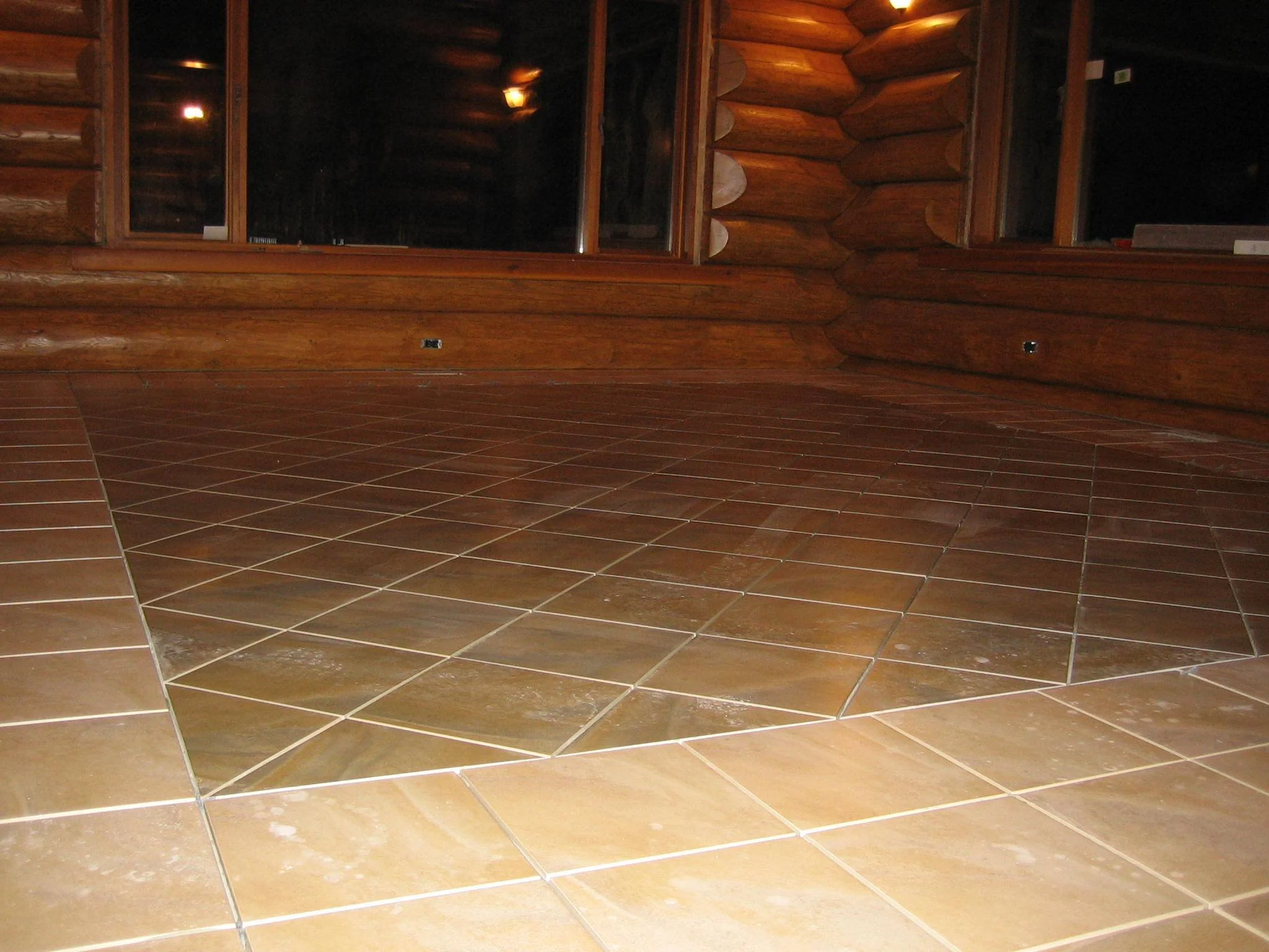 Interior view of a wood cabin with large windows, wooden logs on the walls, and a tiled floor with a diamond pattern.