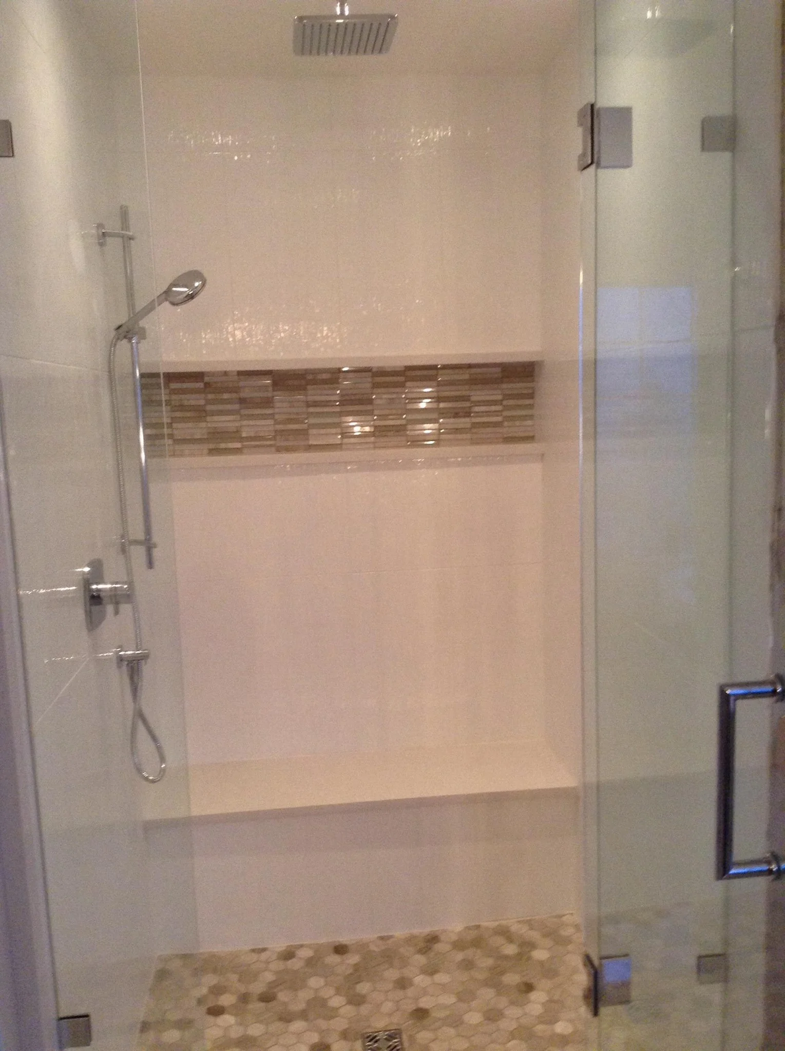 Inside a modern shower with glass doors, featuring a handheld showerhead and a decorative tile strip on the wall.