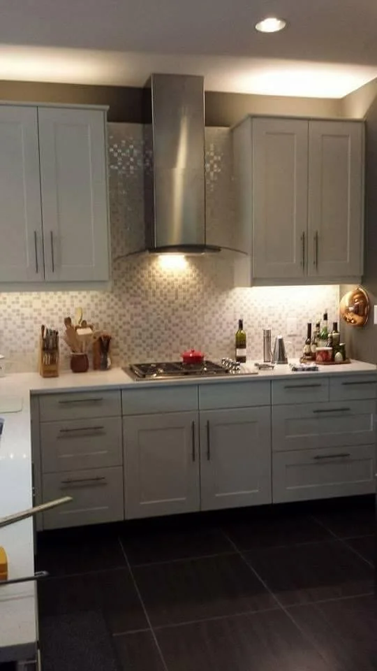 Modern kitchen with gray cabinets, a stainless steel range hood, a gas cooktop, and various bottles and kitchen utensils on the countertop.