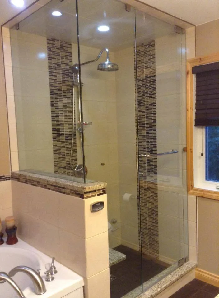 Glass-enclosed shower with a rain shower head, behind a half wall, in a bathroom featuring tile with mosaic accents and a window with a black blind.