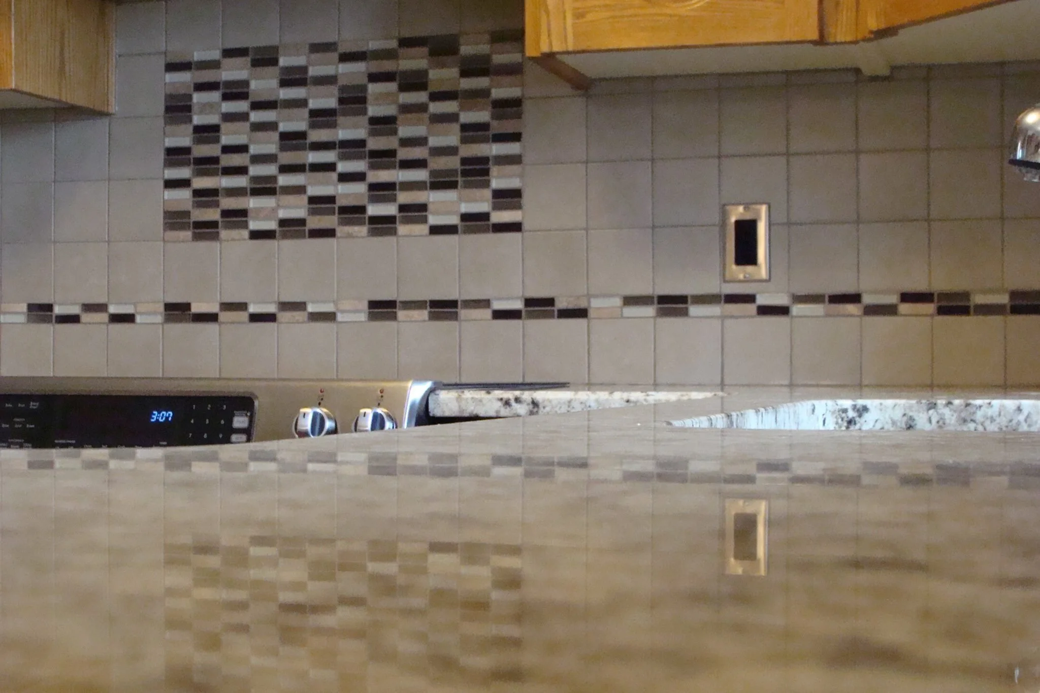 View of a kitchen counter with a granite surface, overlooking a tiled backsplash with a mosaic pattern in brown, beige, and white. Part of a stainless steel stove with digital clock and control knobs is visible in the lower left corner.