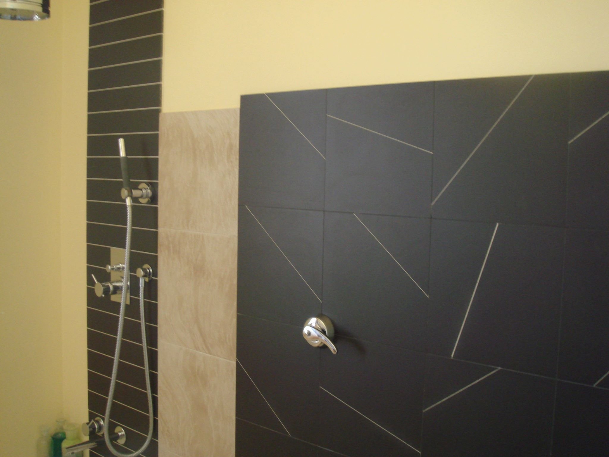 A bathroom shower with a black tiled wall featuring a white geometric pattern, a silver showerhead, and a silver faucet handle. The shower area is separated by a section of beige tile.