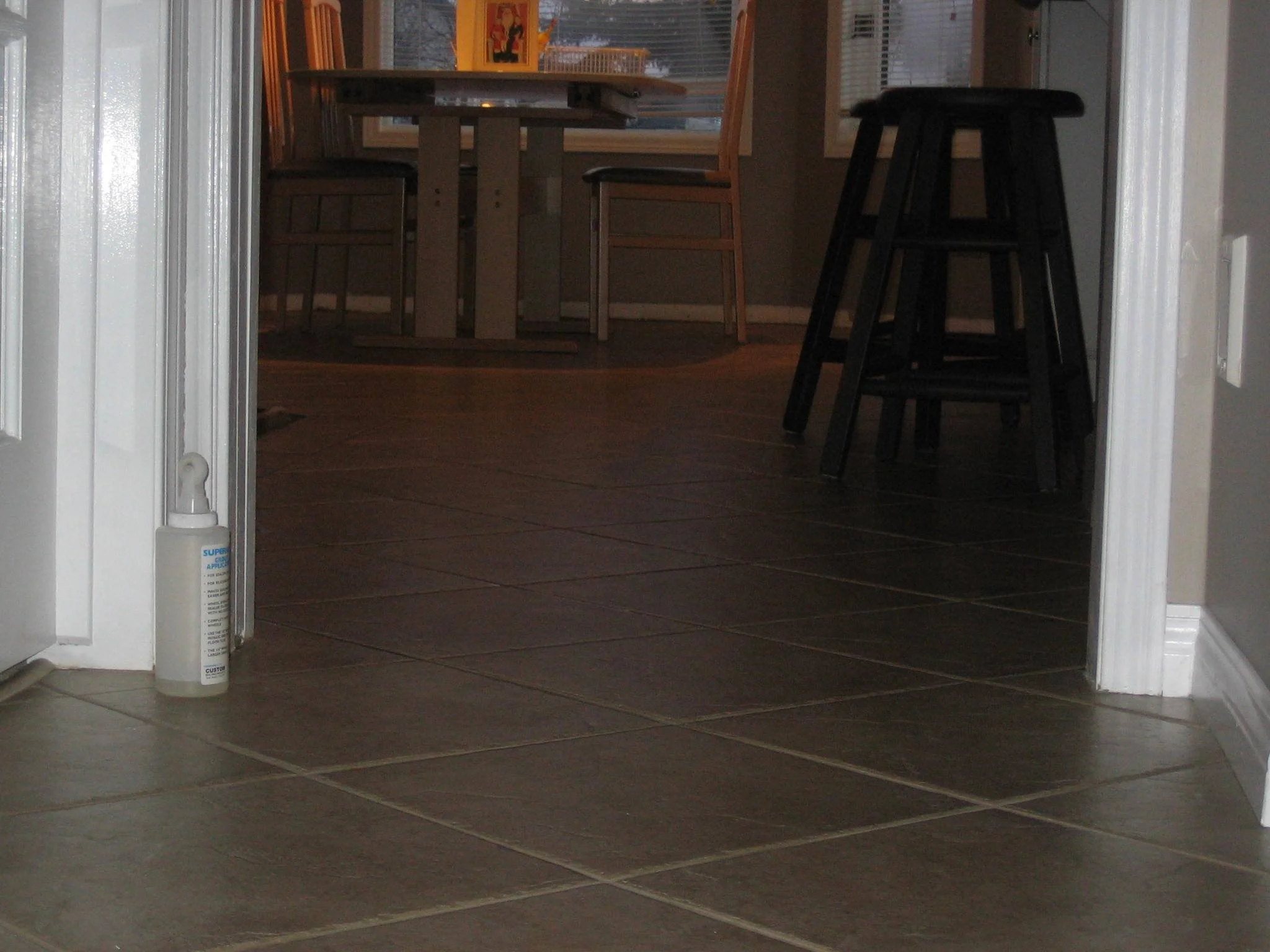 View of a tiled floor leading into a dining area with a wooden dining table, chairs, and bar stools, with a bottle of hand sanitizer near the doorway.
